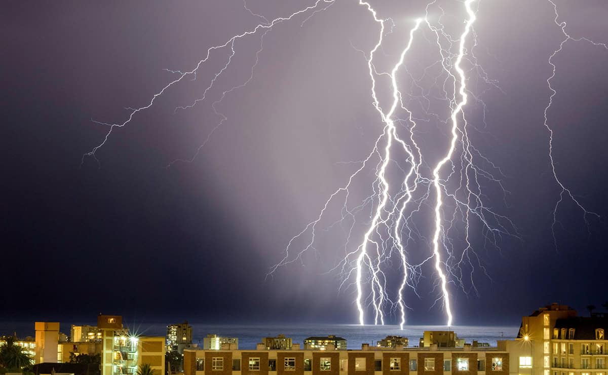 In a dark, cloud-filled sky, several bright bolts of lightening strike a body of water just outside a large city.